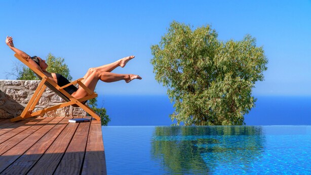 A person reclining on a wooden deck by a clear blue pool with a view of the ocean and a tree. - Villa Asterope, Luxury Retreat by Pleiades (Santorini)