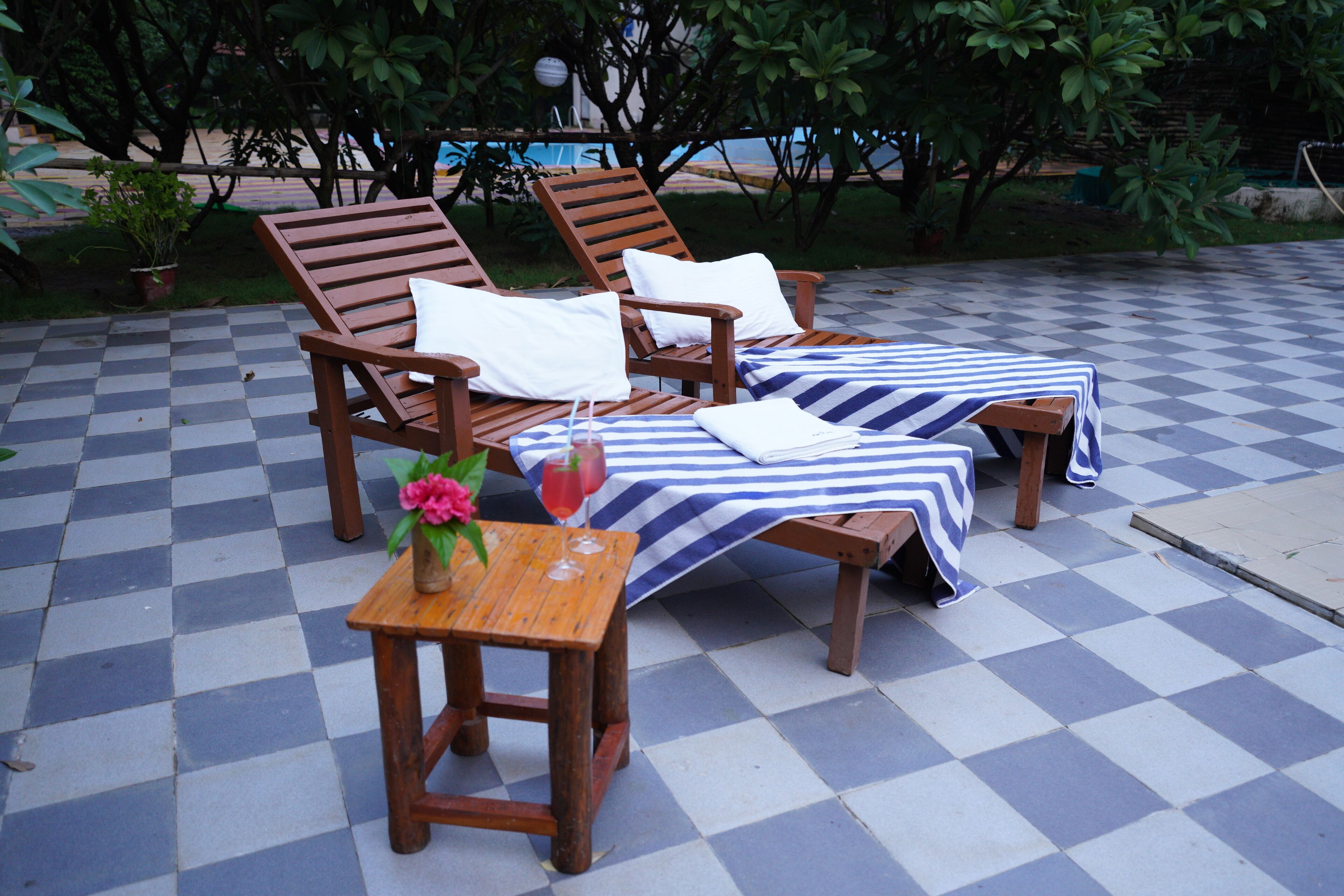 A checkered outdoor seating area with wooden lounge chairs and a small table with a flower vase and drinks.