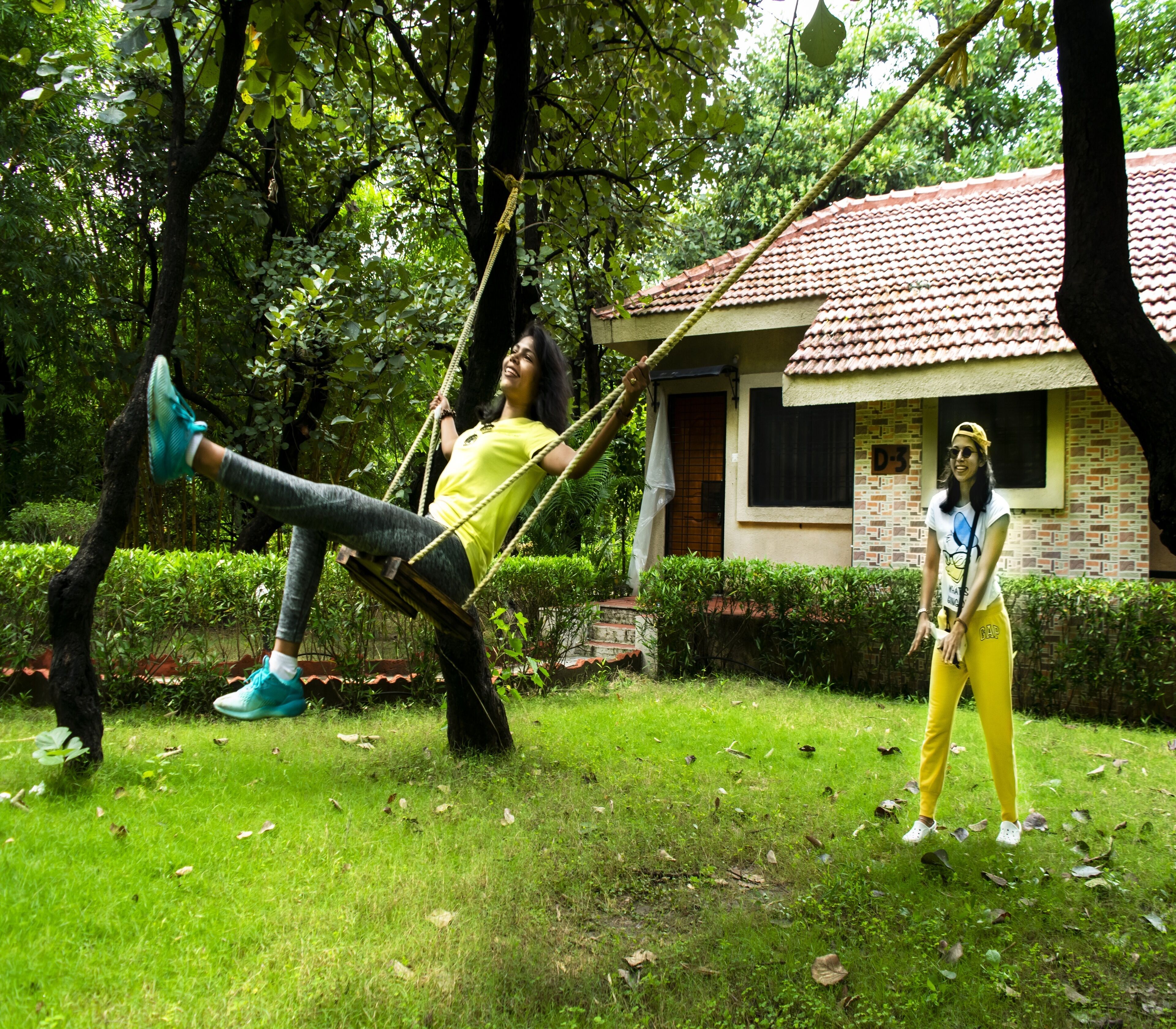 A person on a swing in a grassy area with trees and a building in the background.