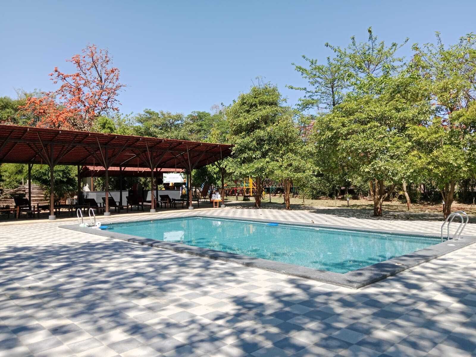A swimming pool with a tiled deck, a covered seating area, and trees in the background.