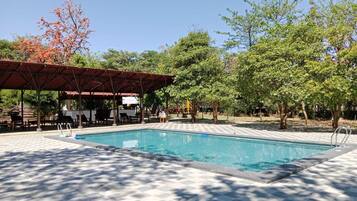 A swimming pool with a tiled deck, a covered seating area, and trees in the background.