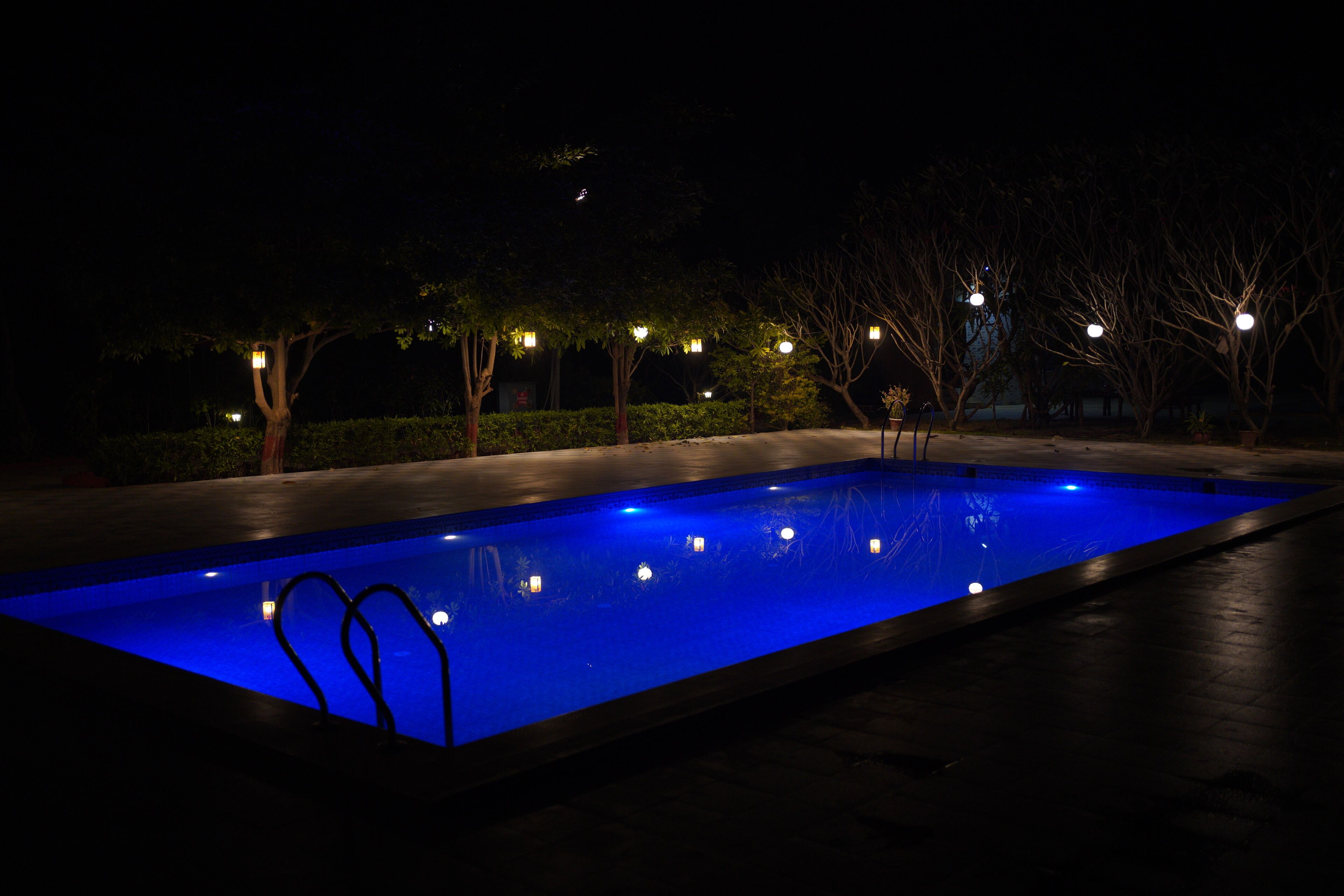 A rectangular swimming pool with a blue illuminated interior, surrounded by trees and illuminated by small lights.