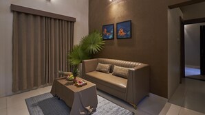 A modern living room with a leather sofa, a coffee table, and a large plant. There are two wall-mounted light fixtures and two framed pictures. - Olive Resorts & Villas (Kurai)