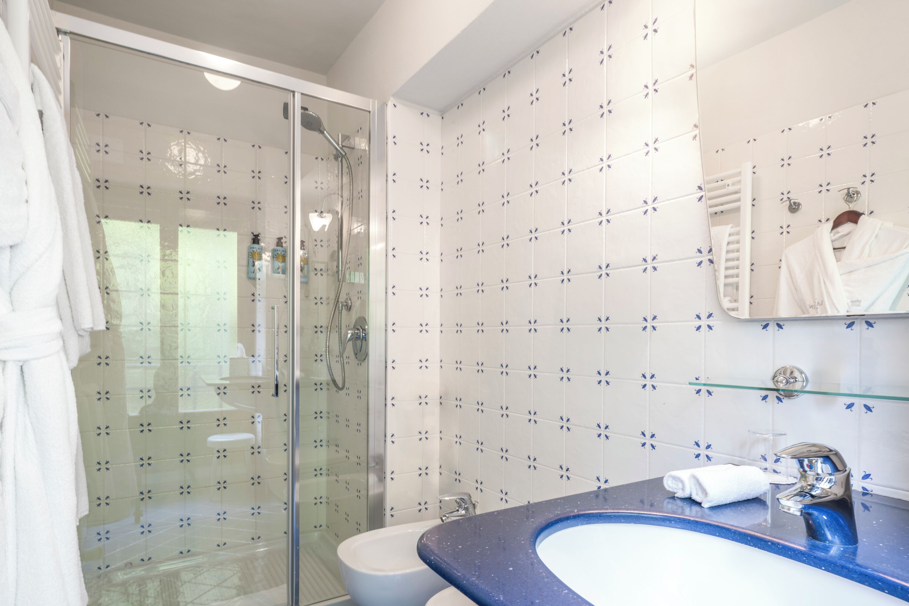 A bathroom with a glass shower enclosure, a white toilet, a sink with a blue countertop, and a mirror with a towel hanging on it.