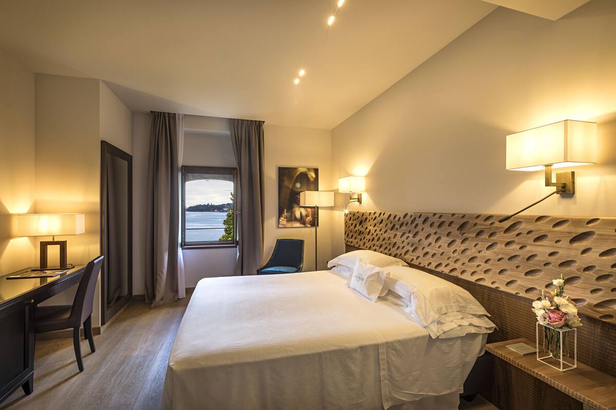 A hotel room with a large bed, a desk with a chair, a window with a view, and two wall-mounted lamps.