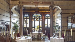 Een statige eetzaal met versierde zuilen, grote ramen en een mozaïekvloer. Tafels zijn gedekt voor een diner, met kaarsen en bloemen.