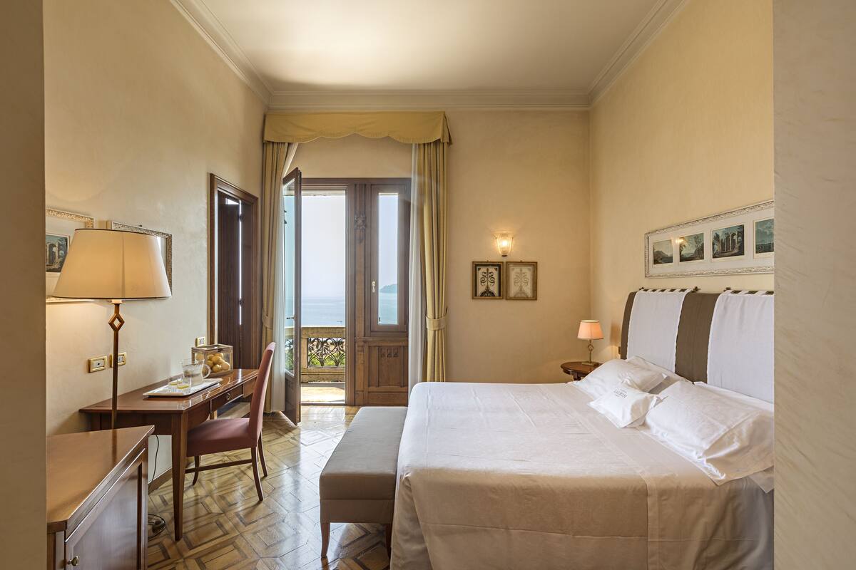 A hotel room with a bed, a desk, a chair, a lamp, and a view of the outdoors.