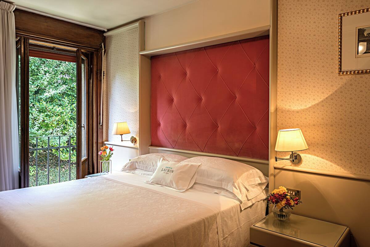A hotel room with a large bed, a red upholstered headboard, two bedside lamps, and a window with curtains.