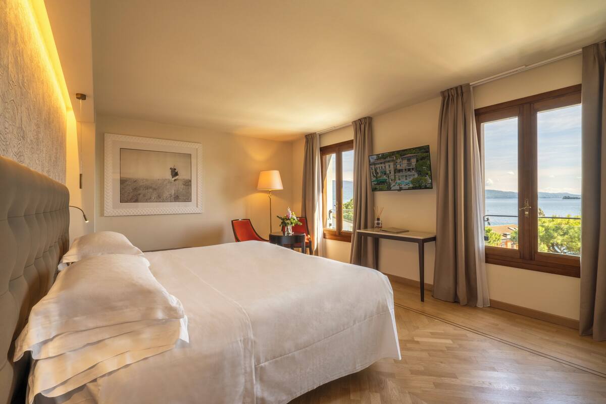 A hotel room with a large bed, a desk, a chair, a lamp, a painting, and a view of the sea.