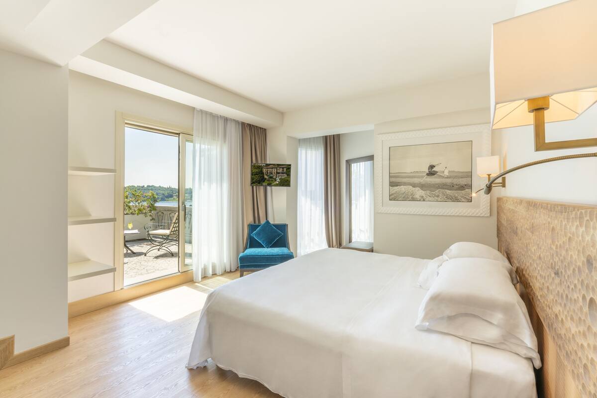 A hotel room with a large bed, a blue chair, a balcony with a view, and a painting on the wall.
