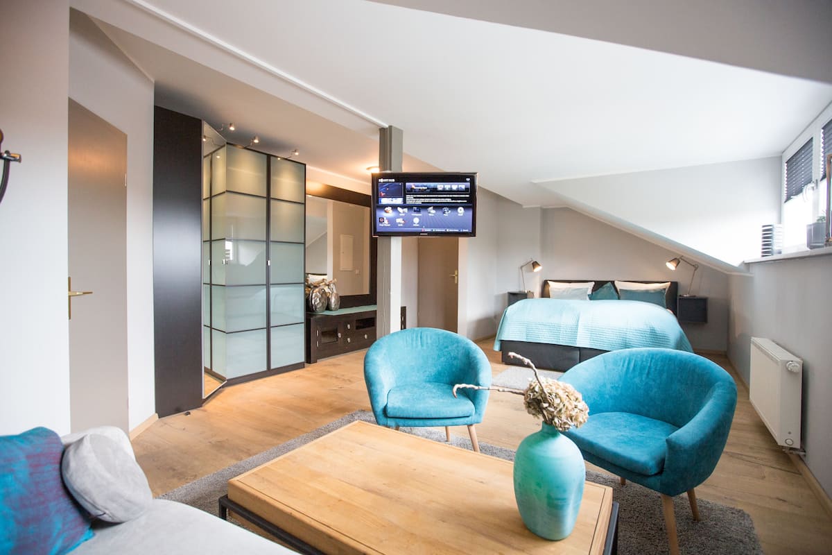 A modern hotel room with a large bed, two blue armchairs, a wooden coffee table, and a flat-screen TV mounted on the wall.