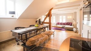 Ein moderner Loft-Wohnraum mit hoher Decke, einem Essbereich mit langem Tisch, einer gemütlichen Sitzecke mit Sofa und einer Küchenzeile mit Herd.