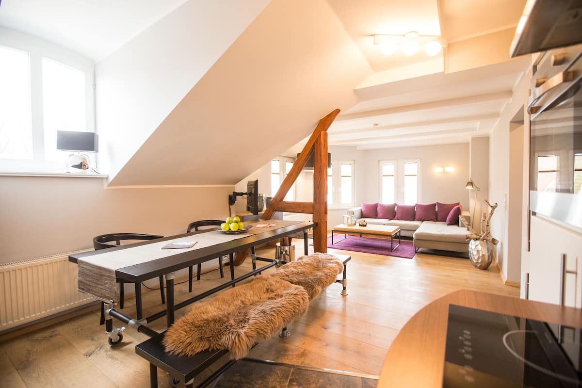 A modern loft-style living space with a high ceiling, a dining area with a long table, a cozy seating area with a sofa, and a kitchen area with a stove.