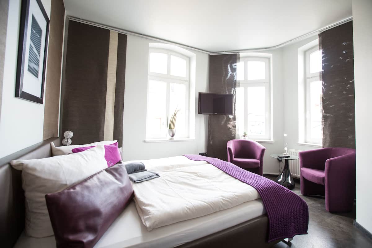 A modern hotel room with a large bed, two armchairs, a small table, a flat-screen TV, and large windows.