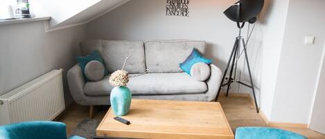 Una sala de estar acogedora con un sofá gris, dos sillones azules, una mesa de centro de madera y un jarrón con flores secas.
