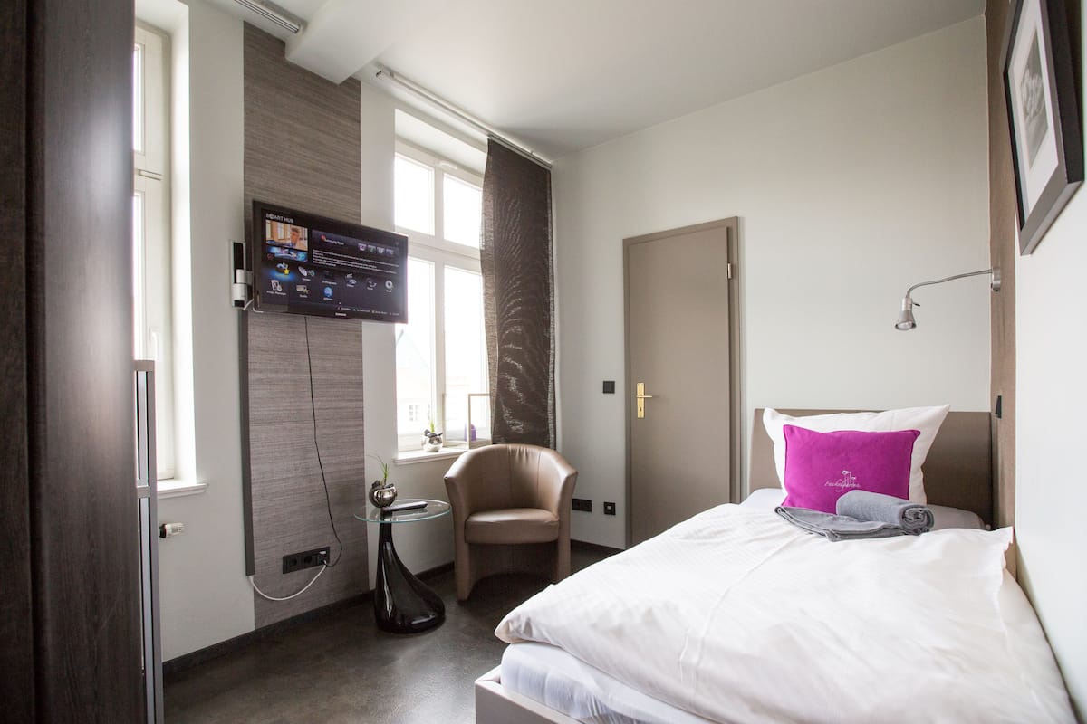 A hotel room with a bed, a chair, a TV mounted on the wall, and a window.