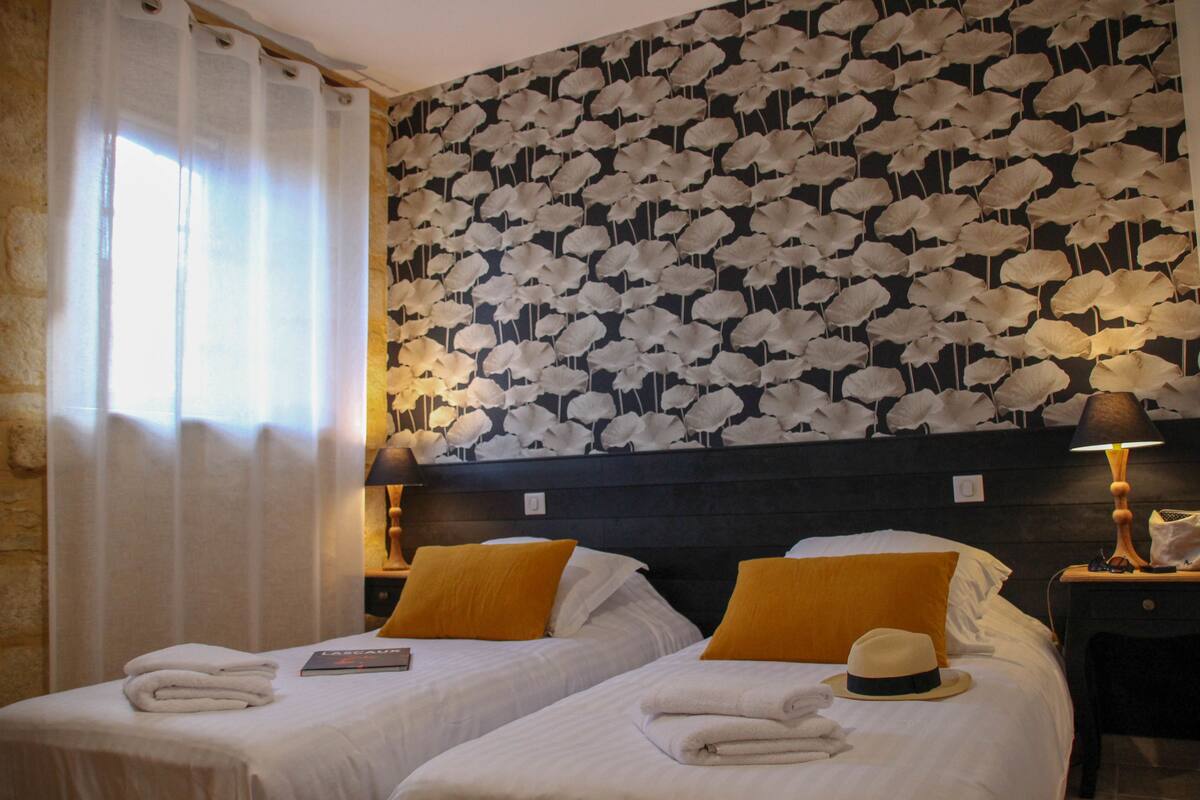 A hotel room with two beds, each with white linens and a yellow pillow. There is a decorative wall with a leaf pattern, a nightstand with a lamp, and a window with curtains.