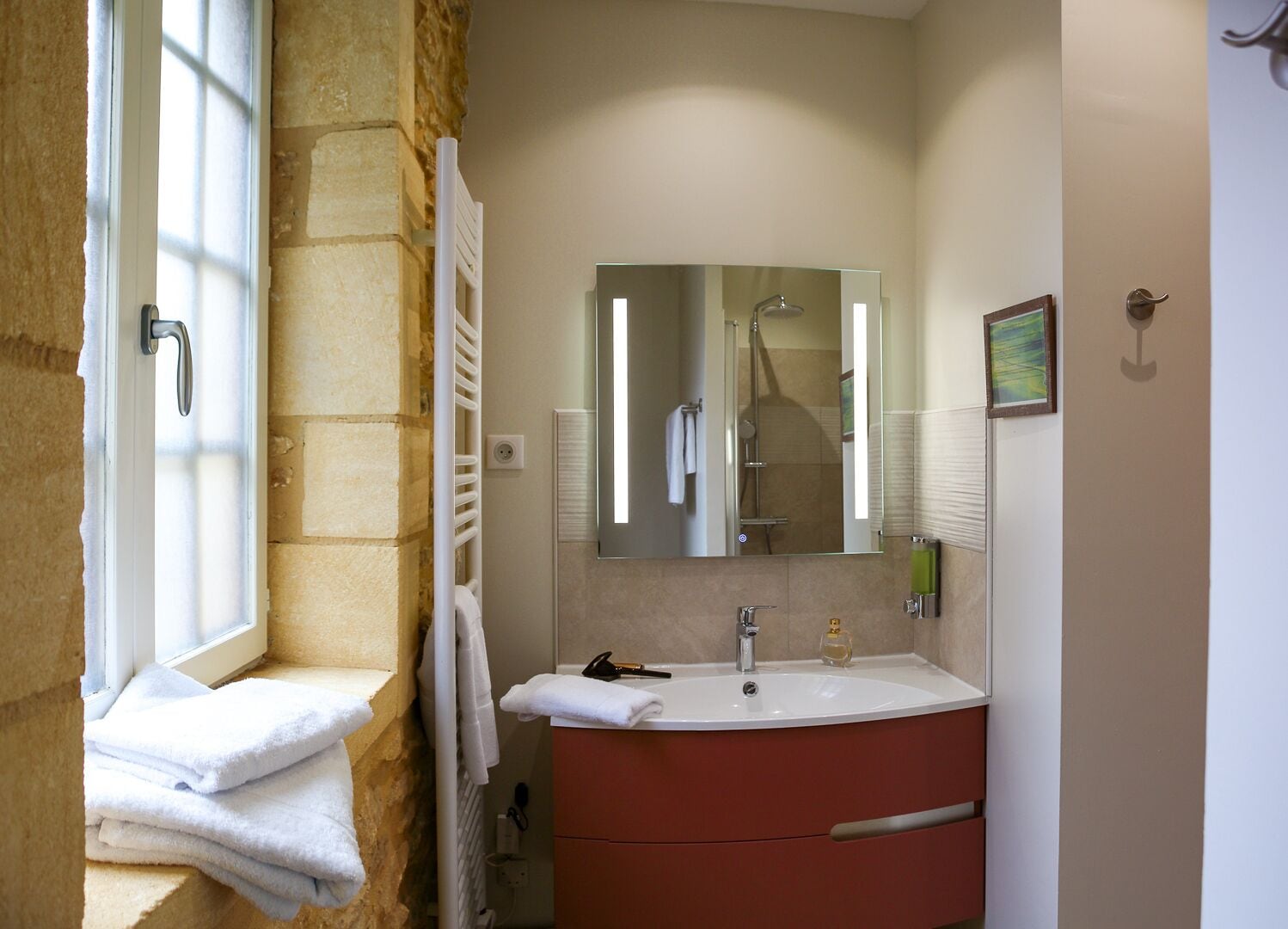 A bathroom with a stone wall, a window with a towel rack, a sink with a mirror, and a shower visible in the reflection.