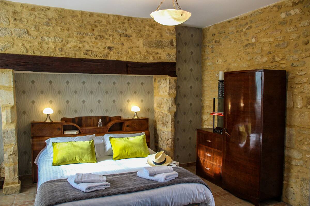 A bedroom with a stone wall, wooden bed frame, two bedside lamps, a wardrobe, and a hat on the bed.