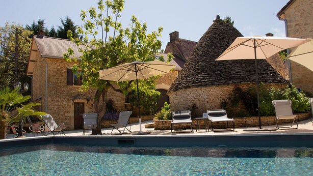 A poolside area with lounge chairs and umbrellas, surrounded by stone buildings and greenery. - Aux Bories de Marquay (Marquay)