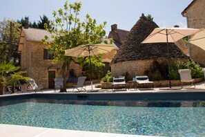 Zona de piscina con sillones de descanso y sombrillas, rodeada de edificios de piedra y vegetación.