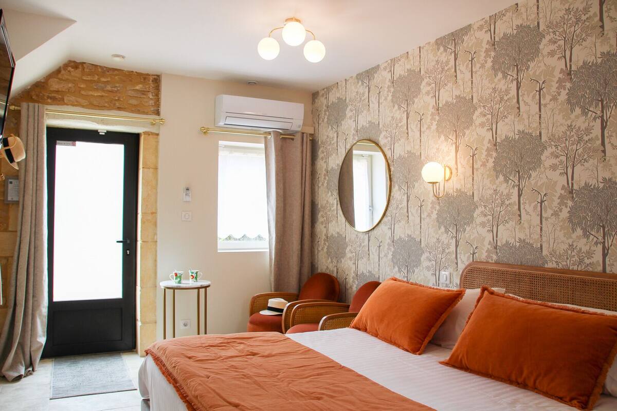 A hotel room with a bed, a round mirror, a small table with a chair, an air conditioning unit, and a door leading outside.