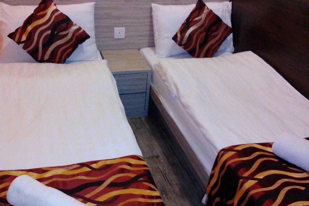 A hotel room with two single beds, each with a patterned bedspread and matching pillows. There is a small bedside table between the beds, and a nightstand with a lamp.