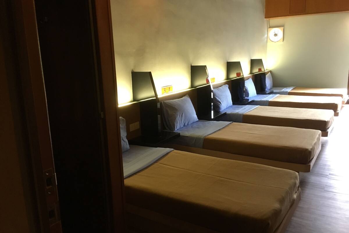 A hotel room with four single beds aligned in a row, each with a headboard and a bedside table.