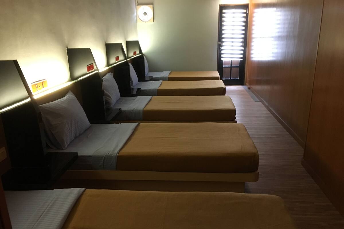 A row of single beds with headboards, each with a small table and a pillow.