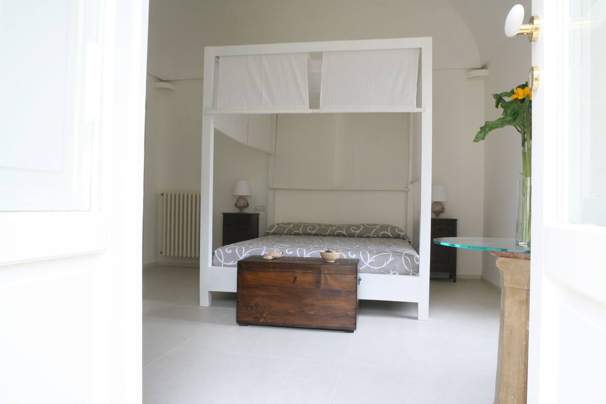 A bedroom with a white four-poster bed, a wooden chest of drawers, a glass table, and a vase with flowers.