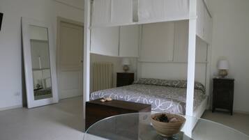 A bedroom with a four-poster bed, a glass coffee table, a wooden side table, a mirror, and a radiator.