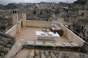 A rooftop terrace with a white table set for dining, a sofa, and several chairs. The terrace overlooks a densely packed urban area with historic buildings.