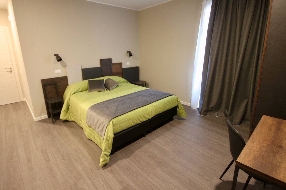A hotel room with a bed, a desk, a chair, a nightstand, and a lamp.