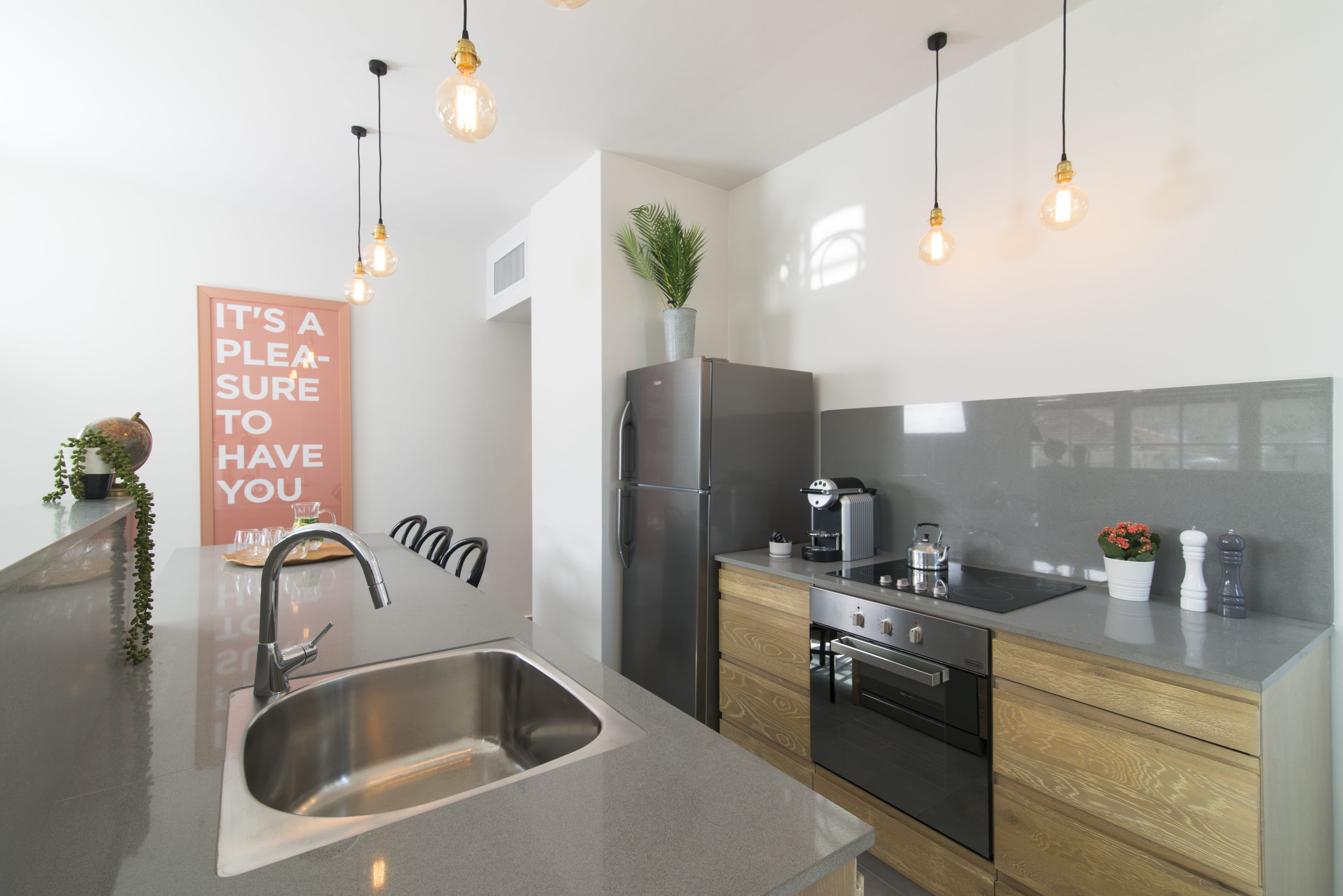 A modern kitchen with a stainless steel sink, a refrigerator, an oven, and a coffee maker. There are hanging lights and a potted plant on the counter.
