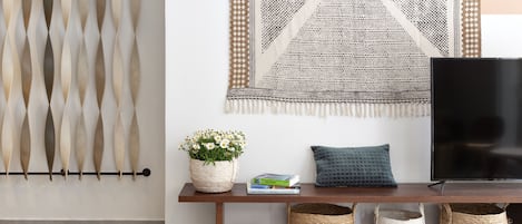 Uma sala de estar moderna com televisão, tapeçaria de tear, mesa de apoio com livros e um vaso de flores, e um varão de cortina fixado na parede.