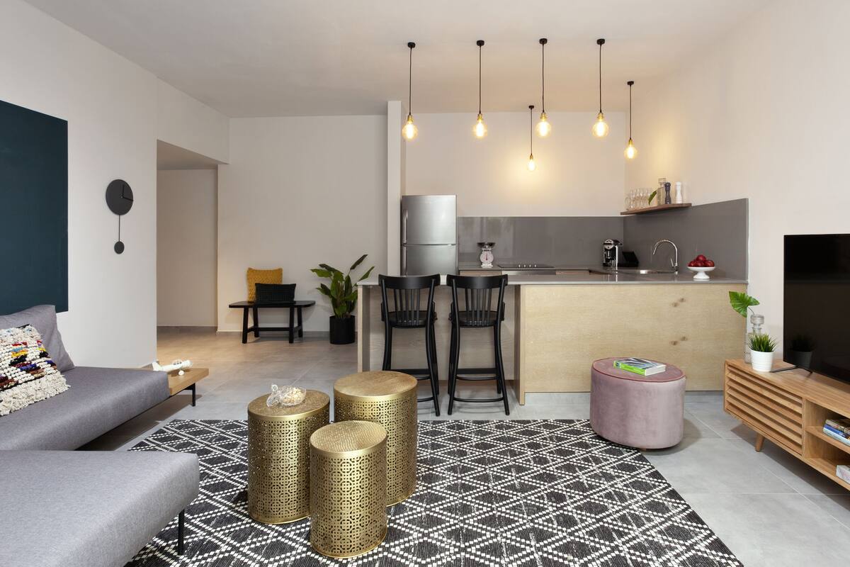 Ein modernes Wohnzimmer mit einem grauen Sofa, einem Couchtisch und einer TV-Konsole mit Büchern. Der Küchenbereich umfasst einen Kühlschrank, einen Herd und eine Spüle.