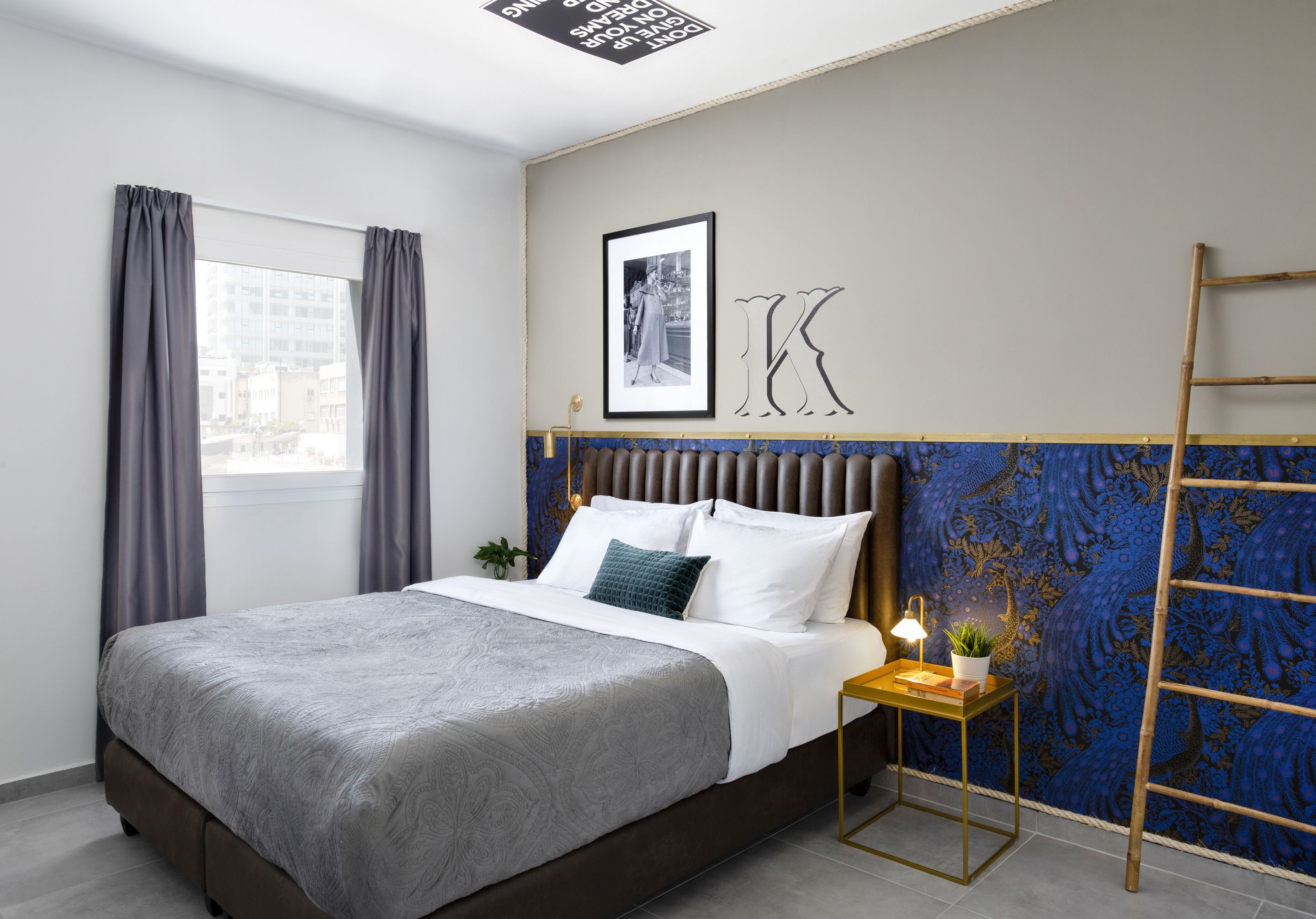 A neatly made bed with a grey blanket and pillows, a bedside table with a lamp and a plant, a framed picture on the wall, and a large letter 'K'.