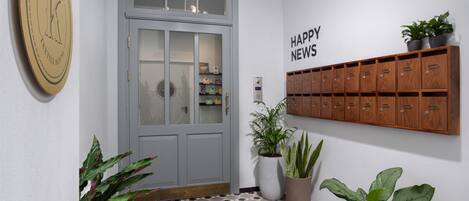 Ein Korridor mit einer grauen Tür, einem Schild mit der Aufschrift 'Happy News', einem Briefschlitz und einer Fußmatte mit der Aufschrift 'Mr. Tel Aviv'.