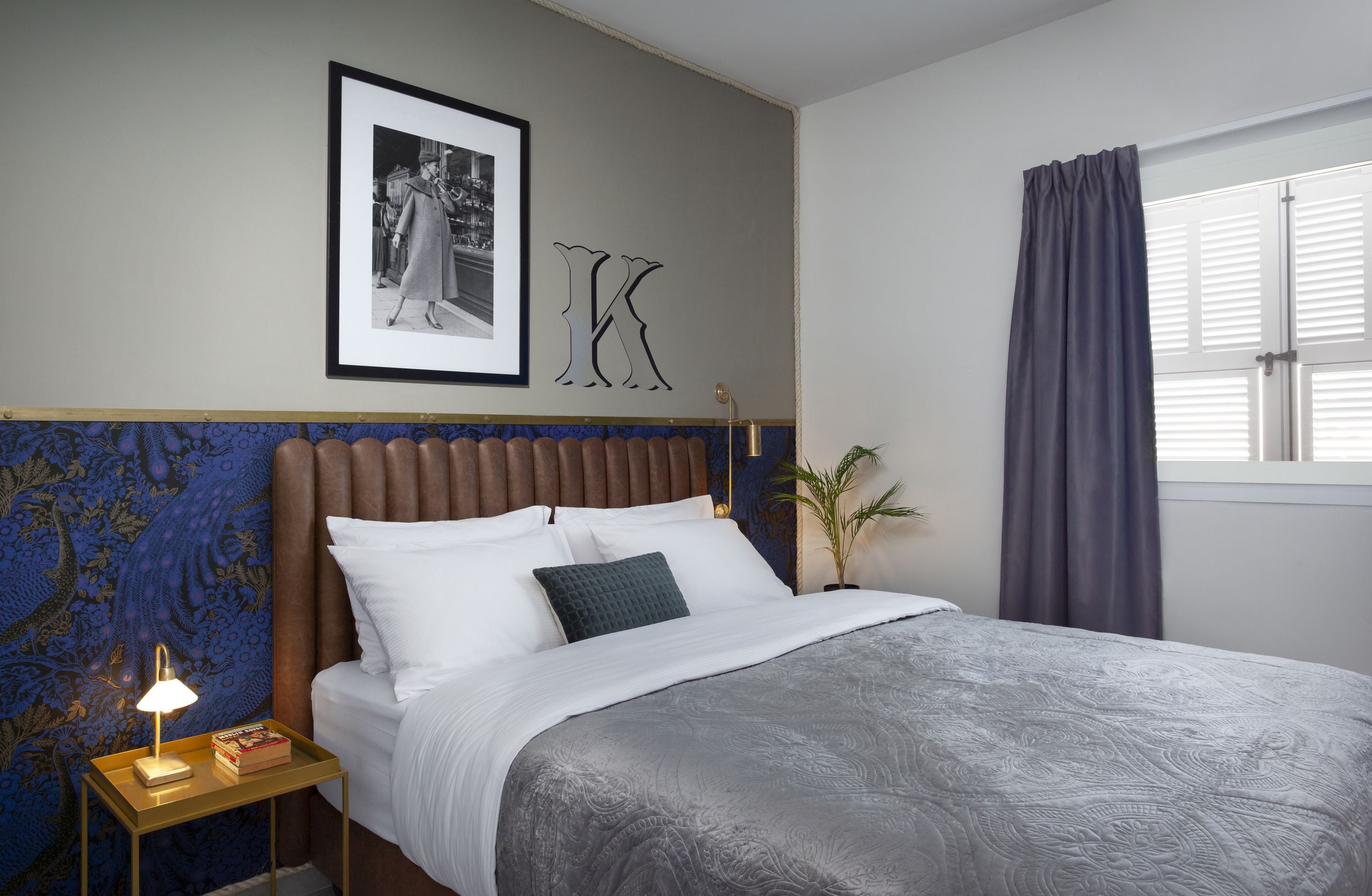 Cama bem arrumada com colcha cinza estampada, uma fotografia em preto e branco emoldurada acima dela, e uma grande letra 'K' na parede.