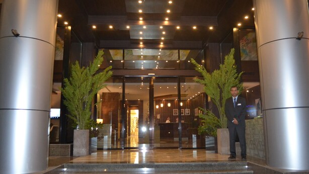 A hotel entrance with a reception desk, two large potted plants, and a man standing by the entrance.