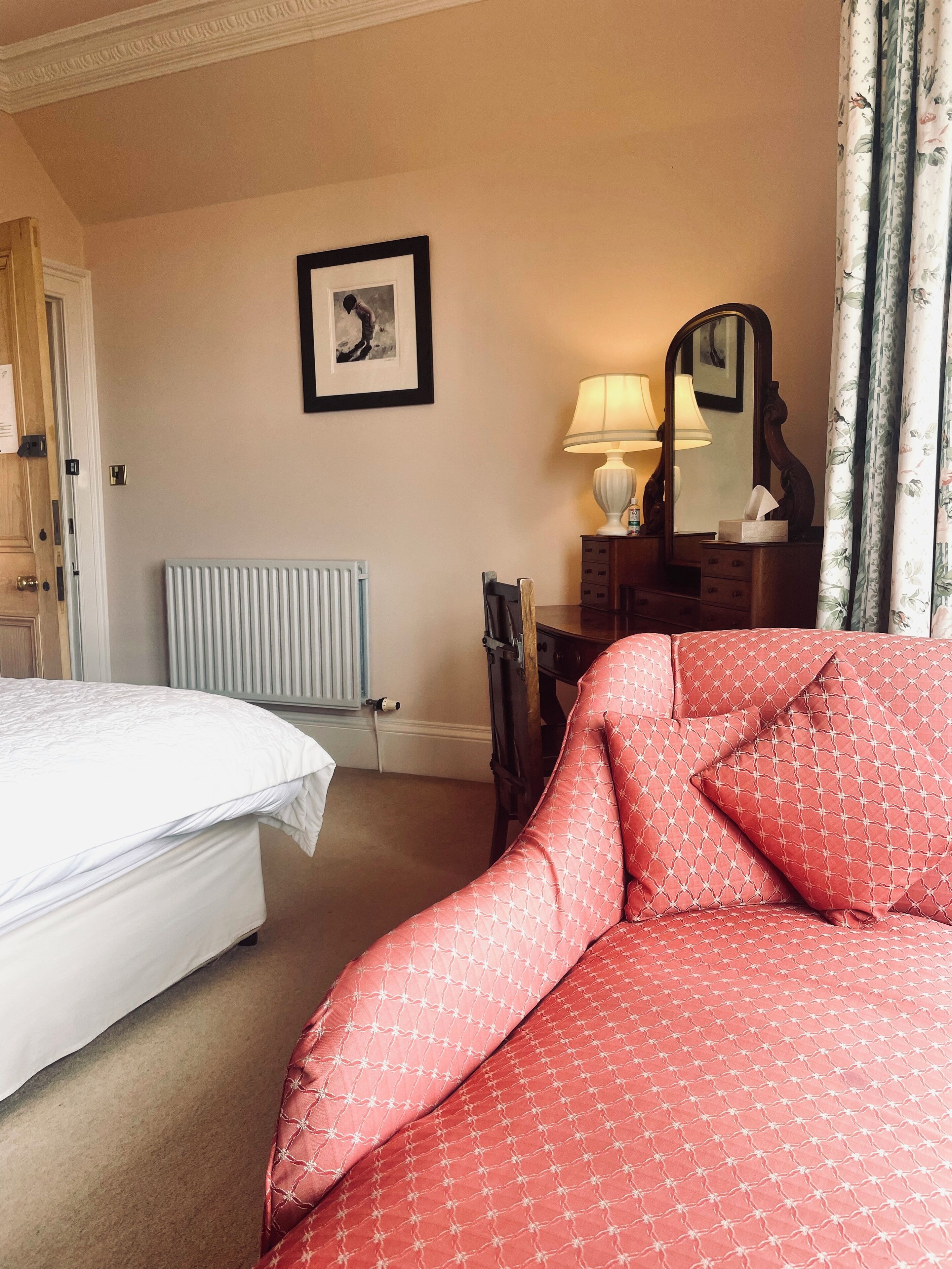 A hotel room with a bed, a desk with a lamp, a mirror, and a radiator.