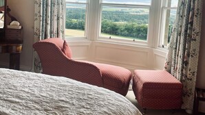 Une chambre avec un grand lit, deux fauteuils rouges à revêtement en tissu, une commode en bois et une vue sur la campagne, visible à travers une grande baie vitrée.