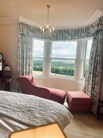 A bedroom with a large bed, two red upholstered chairs, a wooden dresser, and a view of the countryside through a bay window.