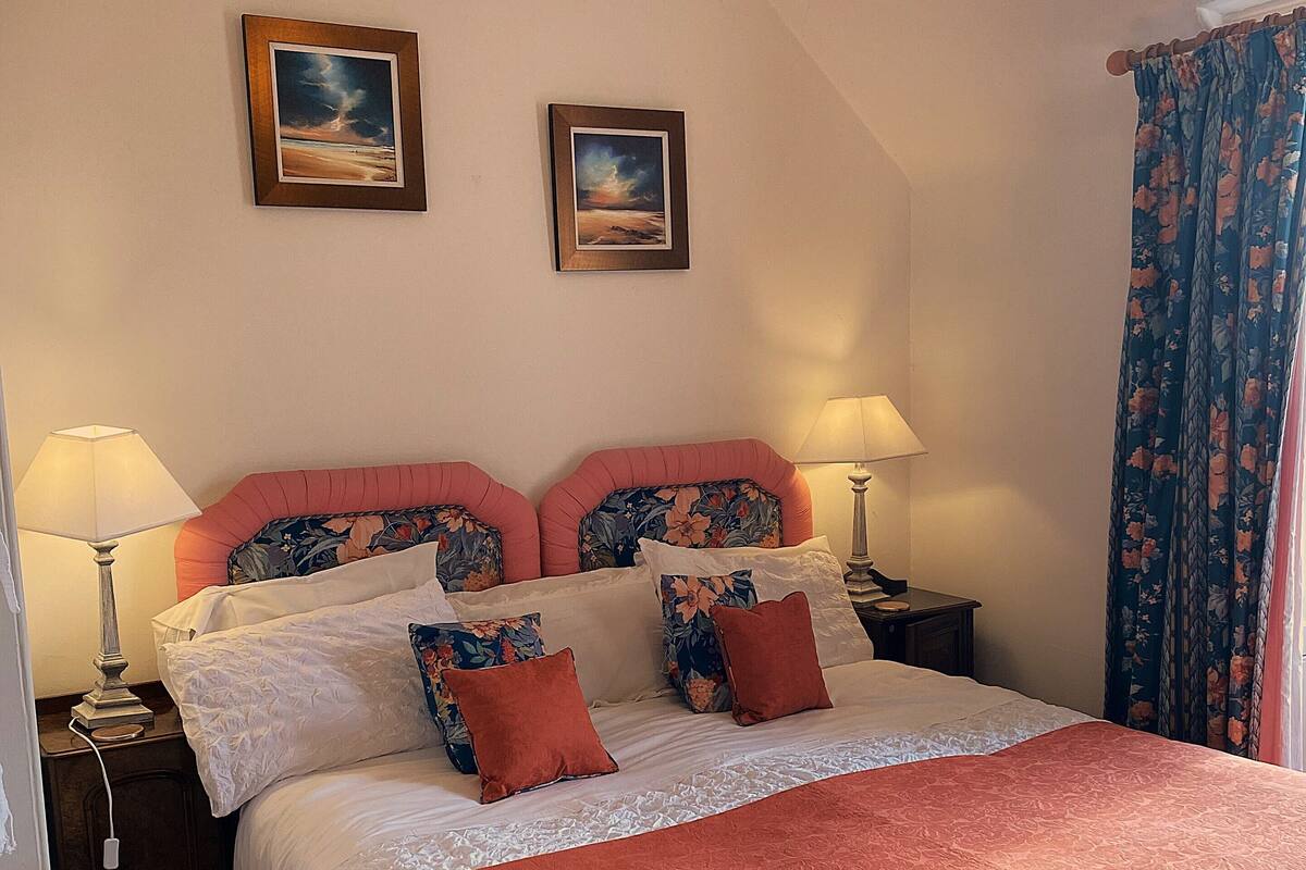 Una cama bien tendida con una colcha roja, dos mesitas de noche con lámparas, y dos cuadros enmarcados en la pared.