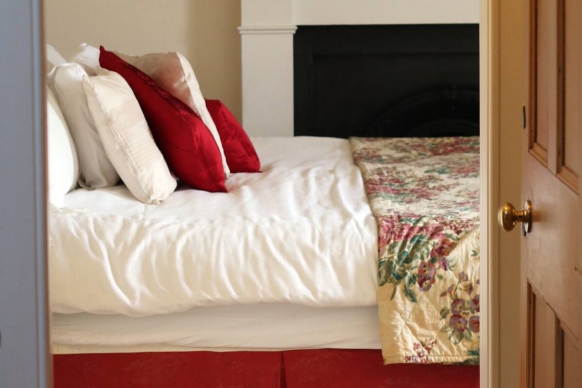 Un dormitorio con una cama, ropa de cama roja y blanca, una colcha con motivos florales, una chimenea y un cuadro enmarcados sobre la repisa.
