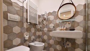 A bathroom with a toilet, bidet, and sink, featuring hexagonal tile walls and a glass shelf above the sink.