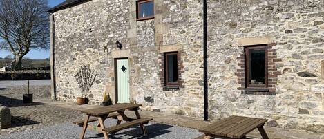 A stone building with a wooden door, two windows, and a picnic table in front.