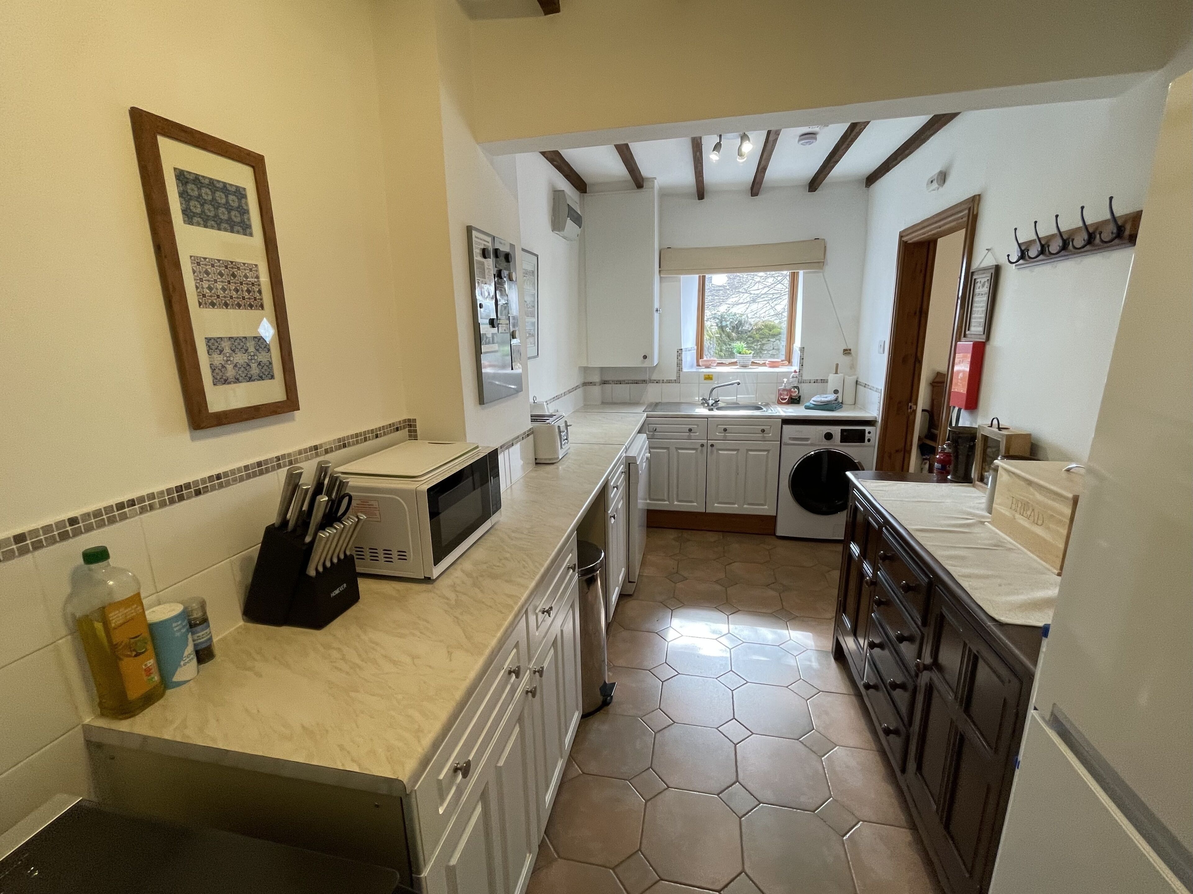 A compact kitchen with a microwave, sink, and washing machine. There are cabinets, a countertop with kitchen utensils, and a window providing natural light.