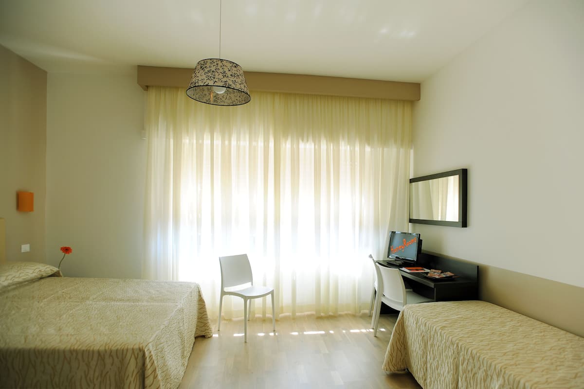 A hotel room with two beds, a desk with a computer, a chair, and a large window with curtains.