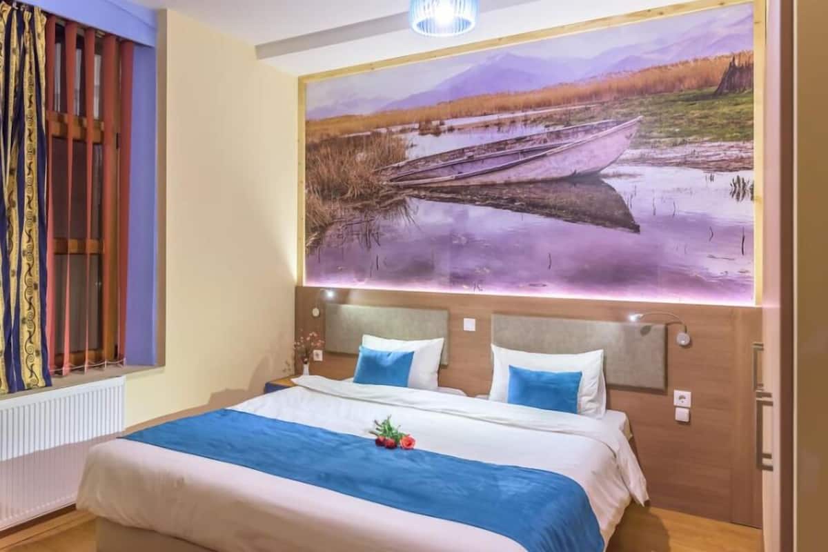 A hotel room with a large bed, two pillows, a nightstand, a window with curtains, and a painting of a landscape on the wall.